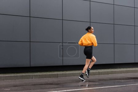 Photo for Full length of brunette african american sportswoman in bike shorts and yellow puffer jacket jogging outside - Royalty Free Image