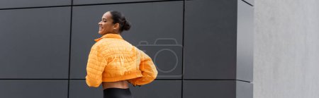 cheerful african american sportswoman in yellow puffer jacket exercising outside, banner 