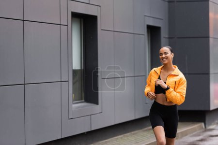 positive african american sportswoman in bike shorts and yellow puffer jacket jogging  outside 