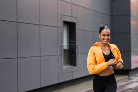 positive african american sportswoman in yellow puffer jacket jogging outside  magic mug #635597428