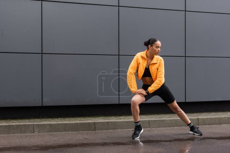 full length of brunette african american sportswoman in bike shorts and yellow puffer jacket exercising outside 
