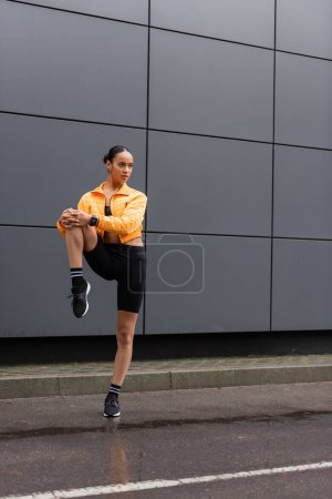 Téléchargez les photos : Full length of brunette african american sportswoman in bike shorts and yellow puffer jacket working out outside - en image libre de droit