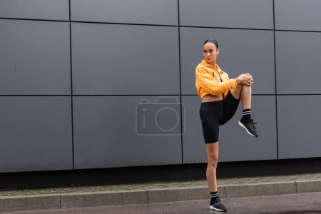 Photo for Full length of brunette african american sportswoman in bike shorts and yellow puffer jacket working out near building - Royalty Free Image