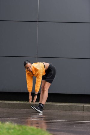 full length of african american sportswoman in bike shorts and yellow puffer jacket stretching near grey building 