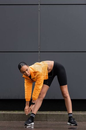 Photo pour Full length of sportive african american woman in bike shorts and puffer jacket stretching near grey building - image libre de droit