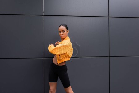 sportive african american woman in bike shorts and yellow puffer jacket training near grey building 