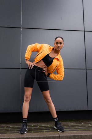 full length of brunette african american sportswoman in bike shorts and yellow puffer jacket exercising with hands on hips 