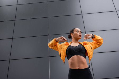 low angle view of brunette african american sportswoman in sports bra and yellow puffer jacket exercising outside 