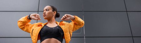 Photo for Low angle view of brunette african american sportswoman in sports bra and yellow puffer jacket exercising outside, banner - Royalty Free Image