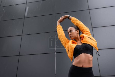 low angle view of african american sportswoman in sports bra and yellow puffer jacket warming up outside 