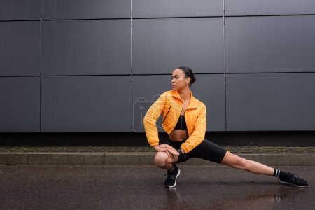 full length of stylish african american sportswoman in bike shorts and yellow puffer jacket stretching near grey building 