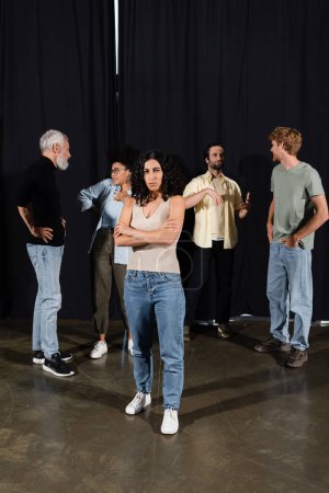 Foto de Multiracial actress standing with crossed arms near interracial theater students and art director talking on background - Imagen libre de derechos