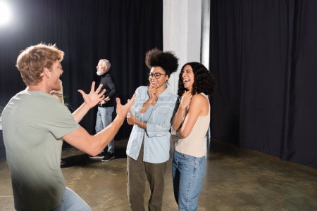 redhead man rehearsing and gesturing near cheerful interracial actresses in theater school