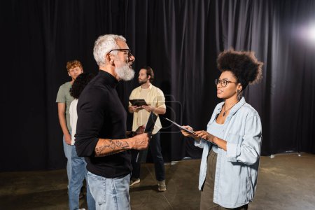 Photo for Tattooed grey haired screenwriter holding scenario near african american woman with clipboard and actors in theater - Royalty Free Image