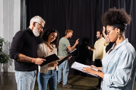 african american woman reading screenplay near bearded art director and troupe rehearsing in theater
