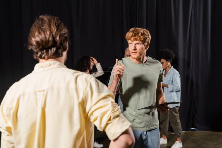Foto de Redhead actor holding clipboard and pointing with finger near blurred man in acting skills studio - Imagen libre de derechos
