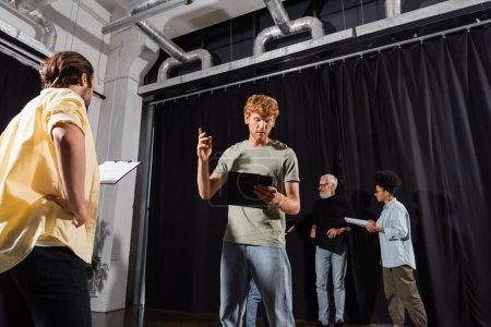 Téléchargez les photos : Redhead man gesturing while looking at screenplay and rehearsing near multiethnic actors in theater - en image libre de droit