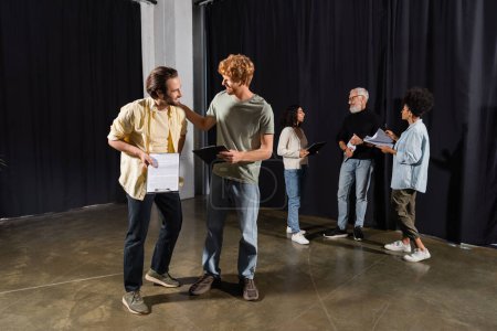 smiling men with scenarios talking in theater school near art director and interracial actors on background