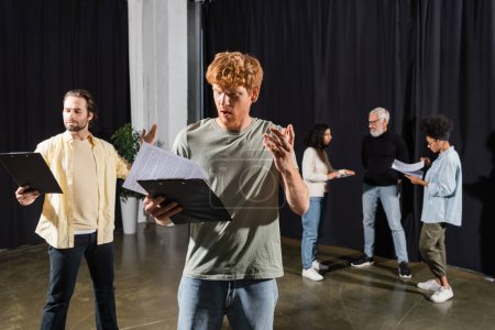 young men holding clipboards with screenplays and gesturing during rehearsing in theater
