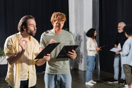 Foto de Brunette and red haired actors holding scenarios while grimacing and gesturing during rehearsal - Imagen libre de derechos
