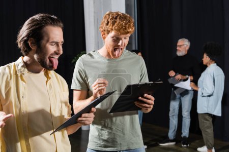 brunette and redhead men sticking out tongues while reading scenarios during rehearsal in theater