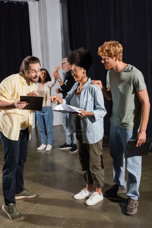 positive man showing clipboard with scenario to interracial actors on stage in theater
