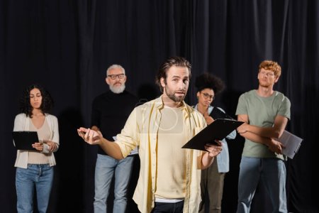 brunette bearded man holding clipboard and looking at camera while rehearsing near multicultural actors and art director in theater