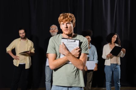 tensed redhead man holding scenario and looking at camera near multiethnic actors and producer on blurred background