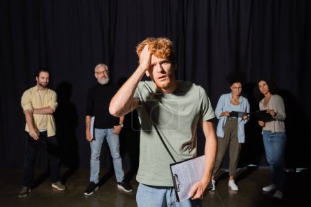 frustrated redhead man holding clipboard and touching head while looking at camera near actors and producer on blurred background