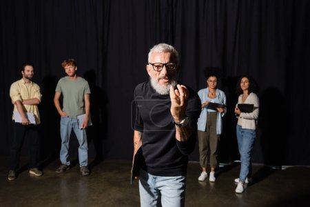 Téléchargez les photos : Bearded art director in eyeglasses looking at camera and gesturing near multiracial students on blurred background - en image libre de droit
