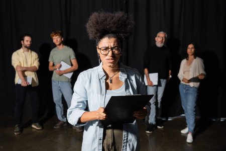Foto de Frowning african american woman holding clipboard and looking at camera near multicultural actors and producer on blurred background - Imagen libre de derechos