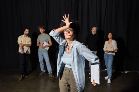 Foto de Emotional african american woman grimacing and gesturing near art director and group of multiethnic artists on blurred background - Imagen libre de derechos