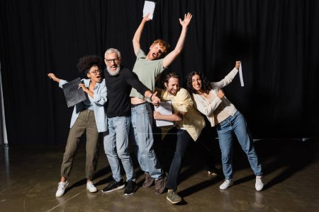 full length of excited interracial actors with bearded art director posing with clipboards on stage of theater