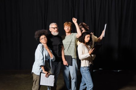 redhead man looking at camera while standing with raised hand near multiethnic actors and bearded producer in theater