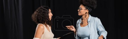 smiling african american woman in eyeglasses pointing with finger while talking to multiracial actress in theater, banner