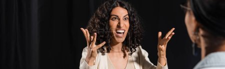 Photo for Multiracial actress with angry face expression grimacing and gesturing near african american woman on blurred foreground, banner - Royalty Free Image