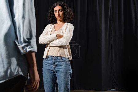 multiracial actress with displeased face expression standing with crossed arms during rehearsal near blurred african american woman 