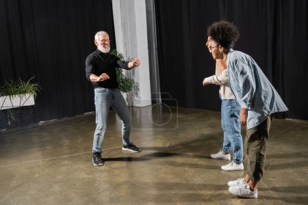 Téléchargez les photos : Interracial actresses laughing near bearded art director posing and gesturing in theater. Translation of tattoo: om, shanti, peace - en image libre de droit