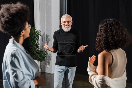 Téléchargez les photos : Cheerful grey haired man pointing with thumb near multiethnic actresses in acting skills school - en image libre de droit