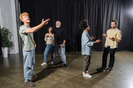 redhead man rehearsing with outstretched hand near art director and multiethnic actors talking on stage in theater