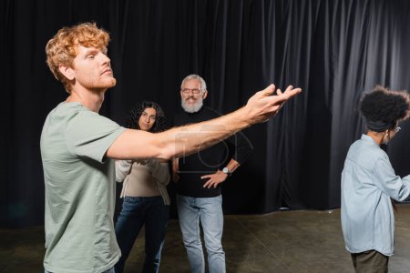 redhead man posing with outstretched hand near interracial women and smiling art director in theater