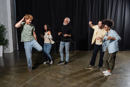 redhead man posing near excited multicultural actors and grey haired producer on stage in acting skills school