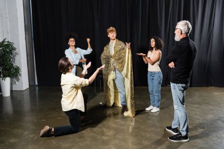 Téléchargez les photos : Young man in king costume standing with outstretched hand near posing actors and grey haired art director - en image libre de droit