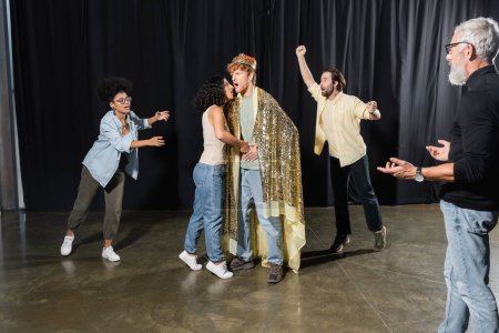 Téléchargez les photos : Bearded producer gesturing near interracial students rehearsing scene of king assassination in theater - en image libre de droit