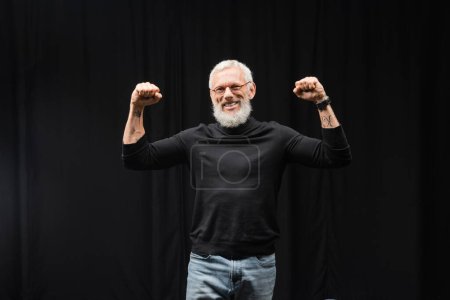 Téléchargez les photos : Excited tattooed actor in turtleneck showing success gesture near black curtain in theater. Translation of tattoo: kanji, danger - en image libre de droit
