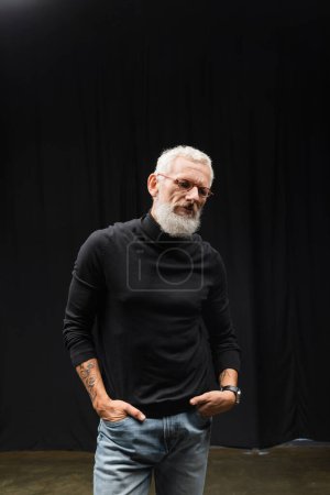 Téléchargez les photos : Mature tattooed actor in eyeglasses and black turtleneck standing with hands in pockets of jeans in theater - en image libre de droit