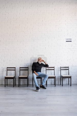 Foto de Bored grey haired man sitting on chair near white wall and waiting for casting - Imagen libre de derechos