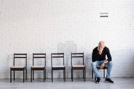 Foto de Full length of pensive man in black turtleneck and jeans waiting for casting on chair near white wall - Imagen libre de derechos