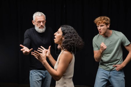 Photo for Multicultural actress rehearsing near gesturing acting skills teacher and emotional redhead man in theater - Royalty Free Image