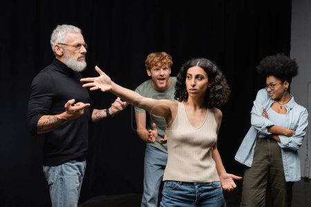 multiracial woman standing with outstretched hand near bearded art director and emotional interracial students in acting school